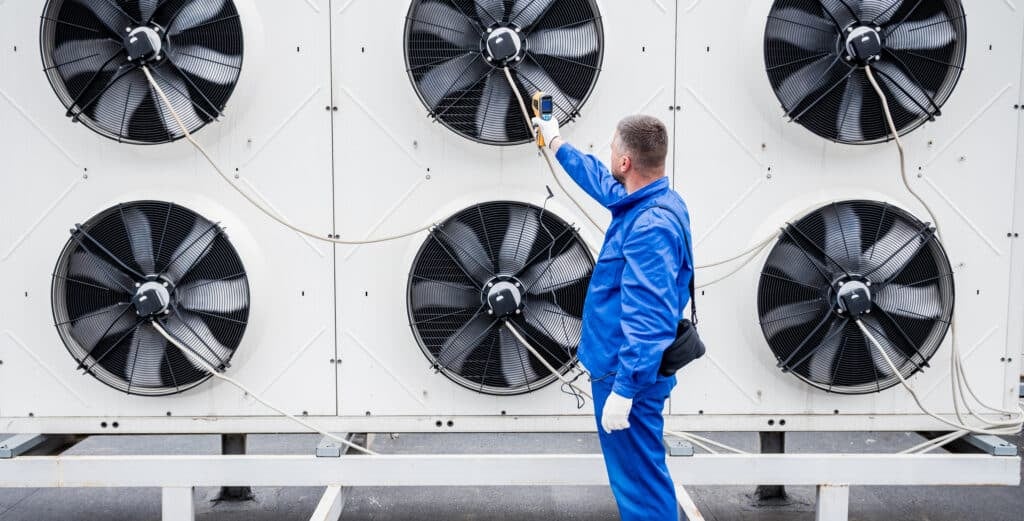 HVAC technician in blue coveralls measuring airflow on a large industrial condenser unit with six fans
